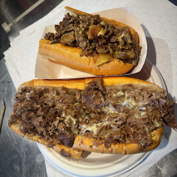 Two Stuff'D Steaks & Hoagies cheesesteaks, one in a paper tray and one on a white plate.