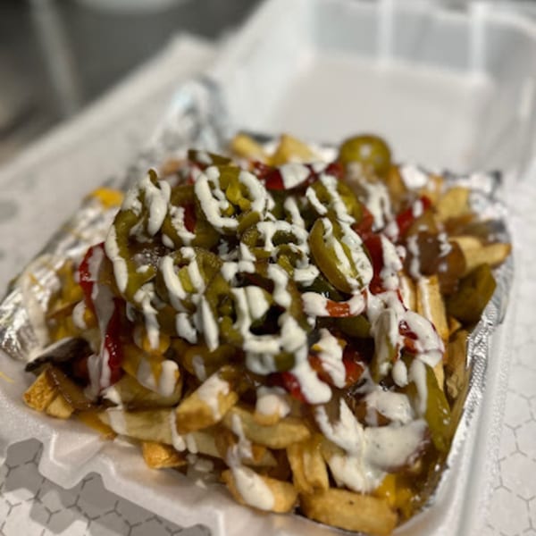 Cheesesteak fries topped with melted cheese, jalapeños, red peppers, and white sauce in a take-out container.