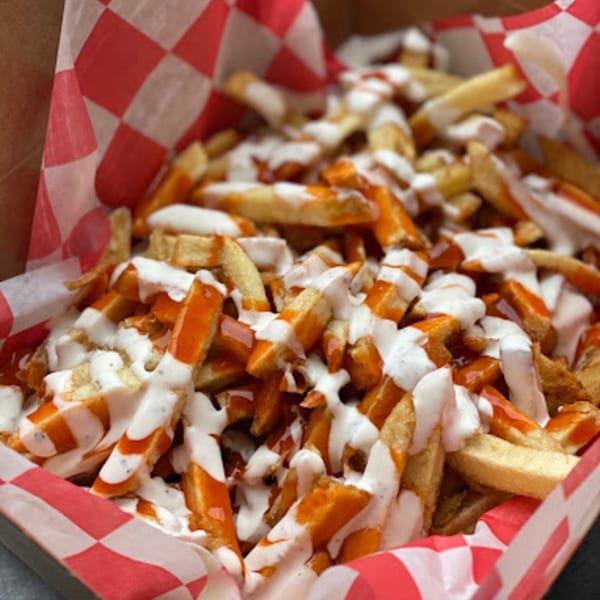 Close-up of golden fries drizzled with white sauce and orange hot sauce in a red and white checkered paper container.