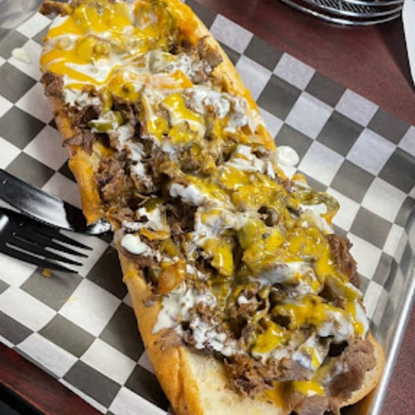 A Stuff'D Steaks & Hoagies cheesesteak, loaded with meat, cheese, and sauce, on a checkered paper-lined tray.